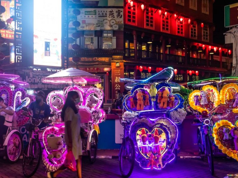 Woman walking across some colourful night carriages on a busy street at Melaka